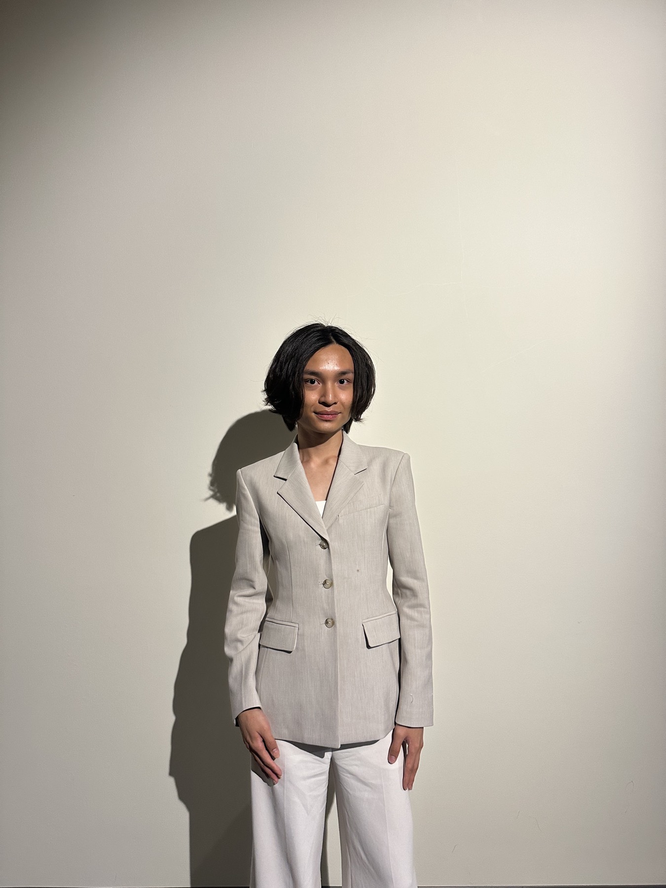Minh Nguyen in a tailored oatmeal blazer and white wide-leg trousers, standing against a cream wall that holds a hard shadow.