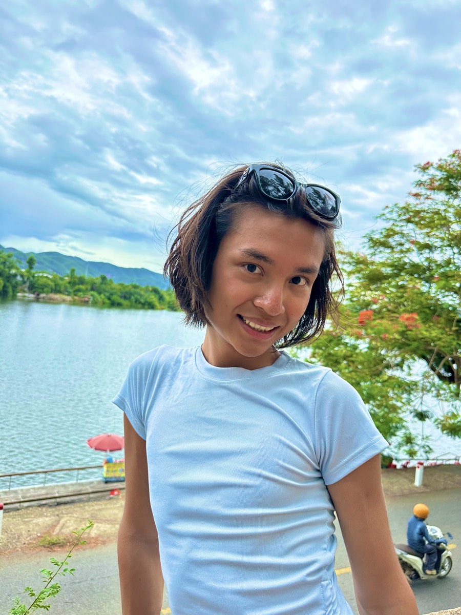 Minh smiling beside a lake with green mountains behind, sunglasses pushed up, in a pale-blue fitted tee.