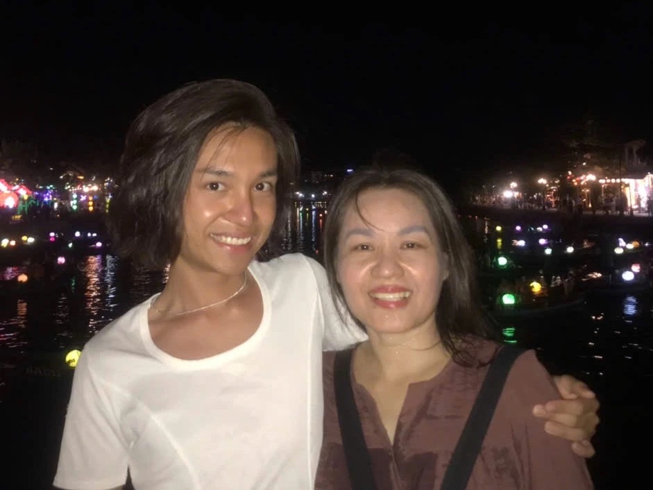 Minh and their mom smiling together at night, Minh's arm around her, with colored lantern lights reflected in the river behind them.